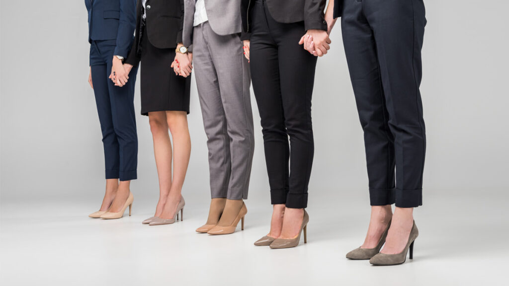 Cropped view of women holding hands and standing in hight heel shoes on grey background